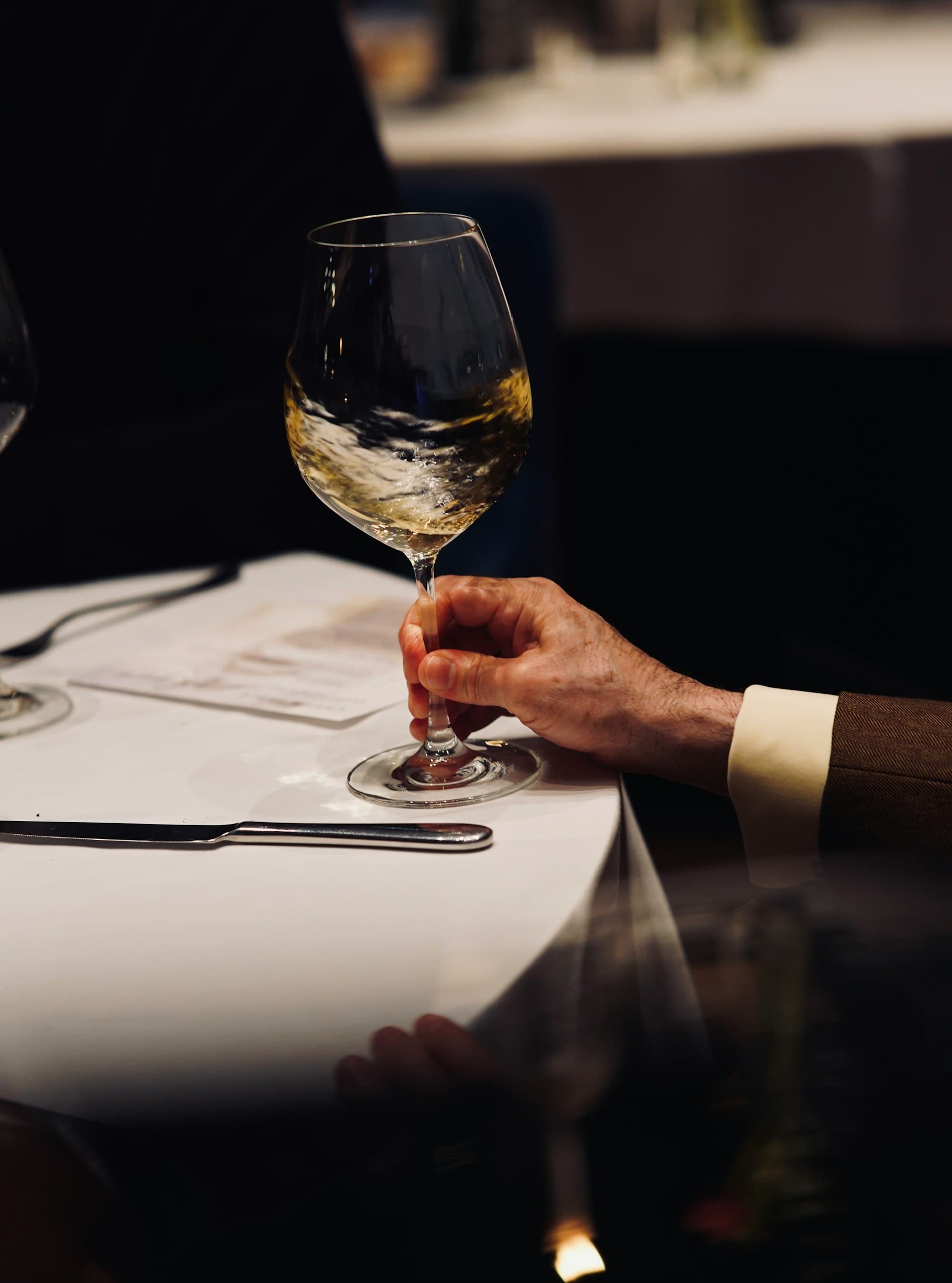 Une main en costume sombre tenant un verre de vin blanc au-dessus d'une nappe blanche